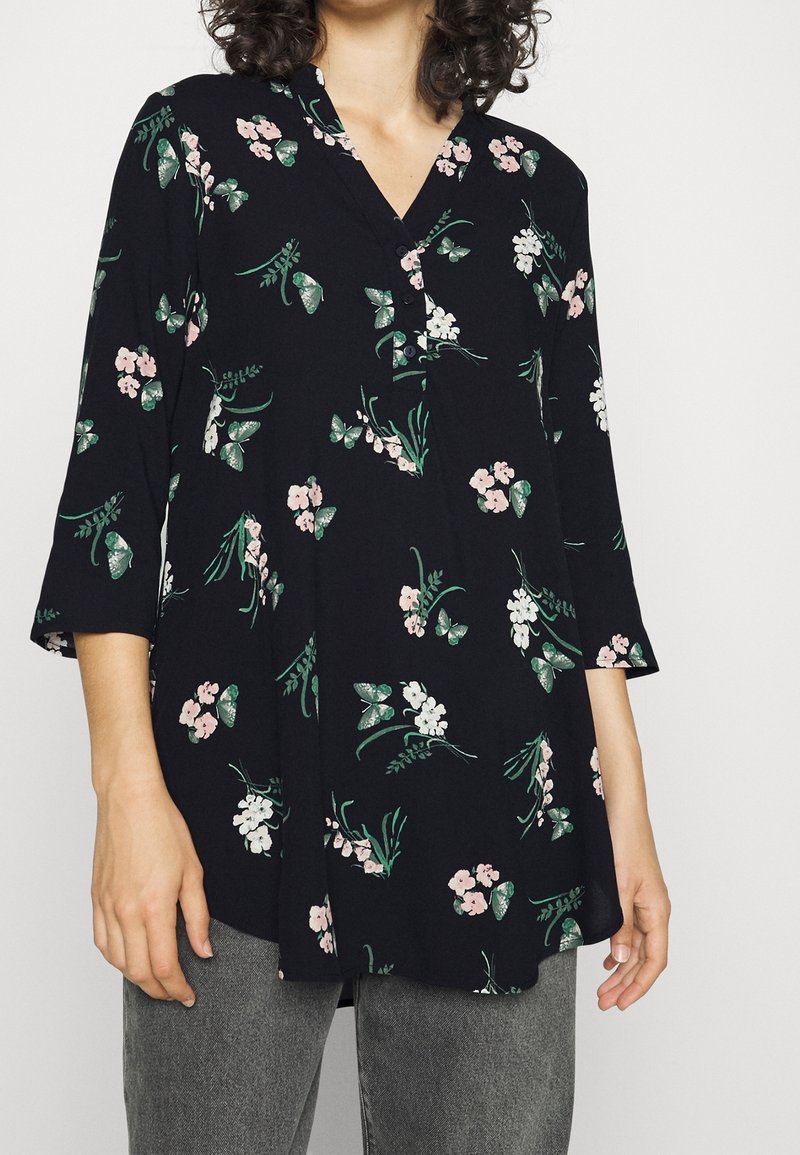 Kvinna iklädd en svart långärmad blus med rosa och vita blommor och fjärilstryck, ihop med grå jeans.