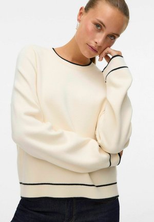 Maglione color crema con dettagli accentuati neri, maniche lunghe, scollo rotondo e tessuto morbido, abbinato a jeans scuri.