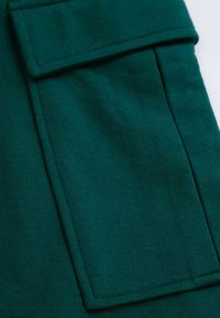 Zielona tkanina z dużą kieszenią z klapką, gładka tekstura i widoczne detale przeszycia.