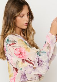Blusa floreale in tessuto trasparente, caratterizzata da colori pastello, maniche a volant e dettagli con paillettes in tutto il design.