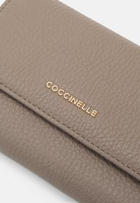 Coccinelle Geldbörse aus taupefarbener Leder mit strukturierter Oberfläche, ausgestattet mit einem goldenen, geprägten Logo und präzisen Nähten entlang der Kanten.