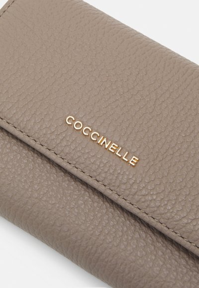 Portefeuille Coccinelle en cuir taupe avec une finition texturée, présentant un logo doré en relief et des coutures précises le long des bords.
