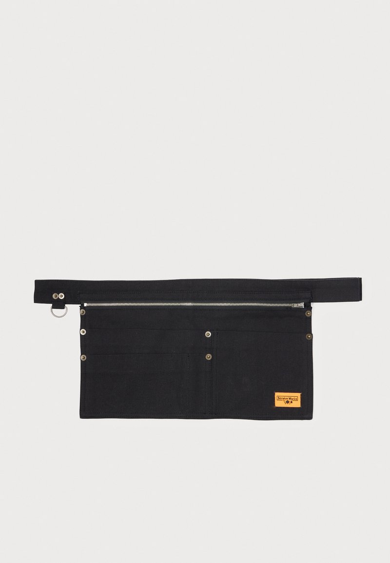 Svart lerretsverktøygj belt med metallglidelås, flere lommer og knappelukkinger, med en liten oransje merkeetikett.