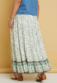 Valkoinen kukkamekko, jossa on vihreitä ja persikkaisia kukkia, kerroksellinen muotoilu ja koristeellinen helma, jossa on monimutkaisia kuvioita. Yhdistetty ruskeisiin kenkiin.