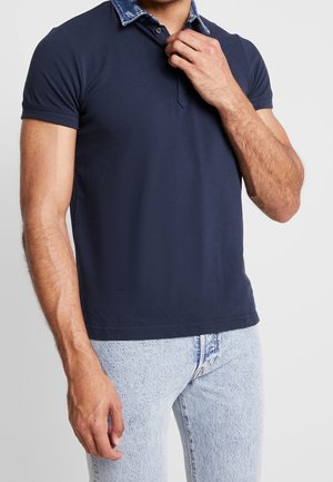 Homme portant un polo bleu marine avec un col en denim et un jean bleu clair, ajustant le col du polo d'une main.