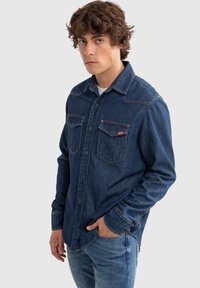 Camisa de mezclilla azul con un cuello clásico, que presenta dos bolsillos en el pecho, costuras visibles y un pequeño acento de logo rojo.