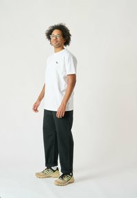 Weißes Baumwoll-T-Shirt mit kleinem schwarzem Logo auf der Brust, kombiniert mit schwarzen Hosen und beigen Sneakern mit grünen Akzenten.