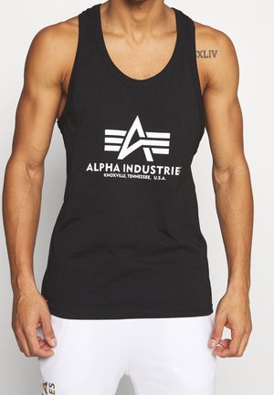 Man wearing black sleeveless tank top with white "Alpha Industries Knoxville, Tennessee, U.S.A." logo and white shorts. Tattoo on right shoulder.