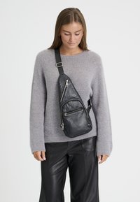 Žena má na sebe šedý sveter a čierne kožené nohavice s čiernou koženou crossbody taškou upevnenou diagonálne cez hruď.