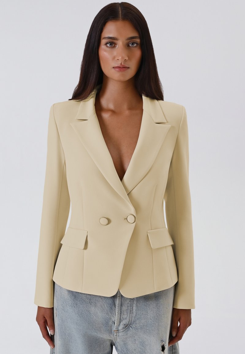 Blazer beige doppiopetto con rever affilati, due tasche frontali e bottoni rotondi, abbinato a jeans chiari strappati.