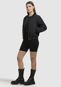 Zwarte gewatteerde bomberjack met een rits aan de voorkant, geribbelde mouwen en zoom. Model draagt zwarte, aansluitende shorts en kniehoge zwarte veterboots.