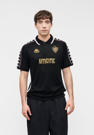 Jeune homme aux cheveux bouclés se tenant face à l'avant, portant un polo noir Kappa avec les inscriptions "AUTHENTIC" et des logos dorés.