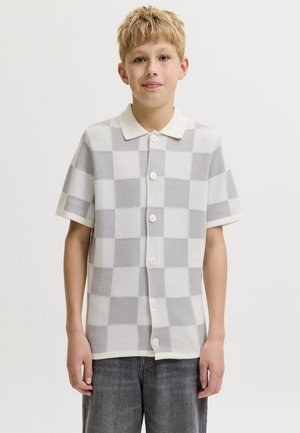 Garçon blond debout portant une chemise à manches courtes boutonnée avec un grand motif damier gris et blanc et un pantalon foncé.