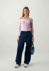 Top rosa decotado com mangas longas, combinado com jeans azuis de perna larga. Uma pequena bolsa cinza é segurada em uma das mãos. Ténis brancos completam o visual.