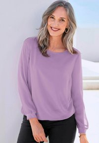 Lavendelfarbene Langarmbluse aus leichtem Stoff mit Rundhalsausschnitt und lässiger Passform, kombiniert mit schwarzen Hosen und dezenten Akzenten.