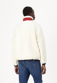 Giacca in pile color crema con alto colletto rosso, orlo elasticizzato blu navy e dettagli a coste sui polsini, abbinata a jeans in denim blu.