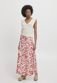 Blusa bianca all'uncinetto con maniche a rouches, abbinata a una gonna lunga maxi con motivo floreale rosso e scarpe con plateau di un beige chiaro.