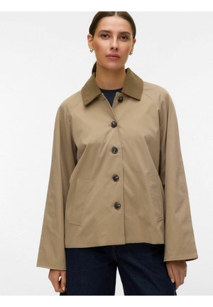 Kobieta w beżowej zapinanej na guziki kurtce z brązowym kołnierzem i ciemnoniebieskich spodniach, stojąca na tle jednolitego jasnego tła.