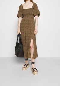 Brun blommig maxi-klänning med puffärmar, med en sidoklaff. Matchad med en svart vävd tote och plattformssandaler med svarta remmar.