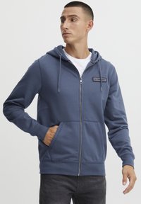 Felpa blu con zip realizzata in tessuto morbido, dotata di tasca frontale, cordoncini regolabili e etichetta cucita sul petto.