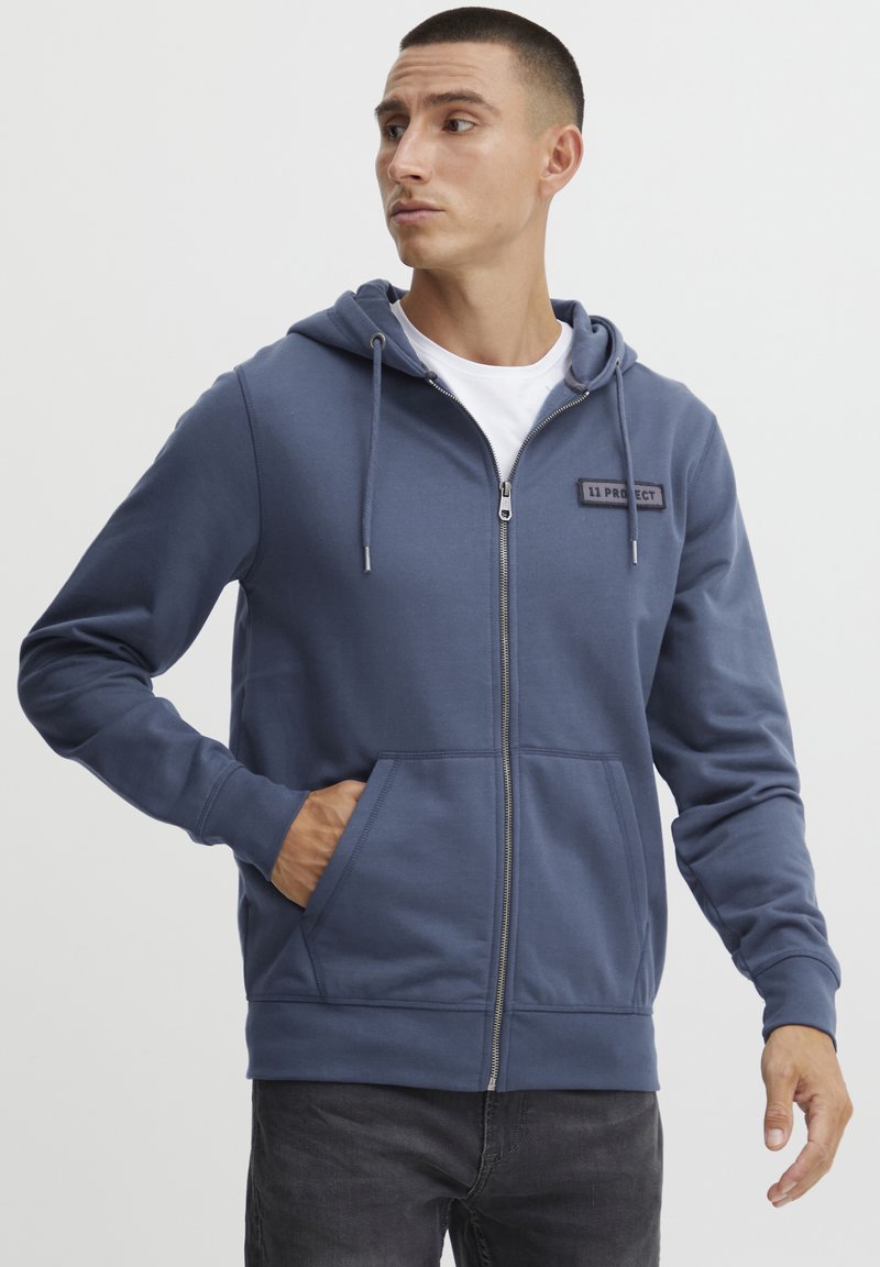 Felpa blu con zip realizzata in tessuto morbido, dotata di tasca frontale, cordoncini regolabili e etichetta cucita sul petto.