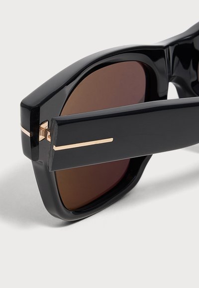 Gros plan sur des lunettes de soleil rectangulaires noires avec des verres marron et un détail d'une fine ligne horizontale dorée sur la branche.