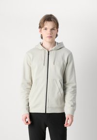 Felpa zip grigia chiara in tessuto morbido. Presenta un cappuccio con cordoncino, tasche frontali e una piccola etichetta con logo sul petto. Zip nera.