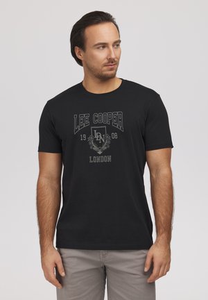 Mężczyzna w czarnej koszulce Lee Cooper London z szarym nadrukowanym logo, patrzy na bok, stoi na jednolitym jasnym tle.