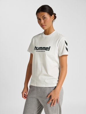 Ung kvinde står, iført en hvid Hummel T-shirt med sort logo og grå joggingbukser mod en ensartet lys baggrund.