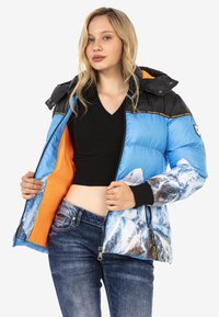 Blauer Pufferjacke mit Bergdruck, schwarzen Schultern, orangefarbener Fütterung und gerippten Bündchen. Verfügt über einen Reißverschluss und ein Logodetail.