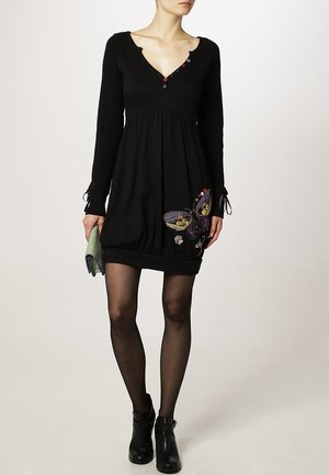 Femme portant une robe noire à manches longues avec un décolleté plongeant et une broderie de papillon sur la jupe, tenant une pochette à motifs et portant des bottines noires.