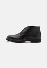 Black leather ankle boots feature a smooth finish, rounded toe, four eyelets for laces, and a subtle emblem on the heel. Flat rubber sole.
