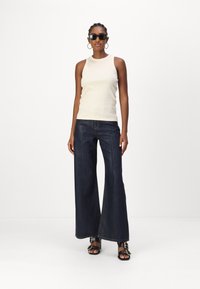 Beige ärmelloses Oberteil mit hohem Kragen, kombiniert mit dunkelblauen weit geschnittenen Jeans mit einer Vordernaht. Schwarze Sandalen mit goldenen Hardware-Details.