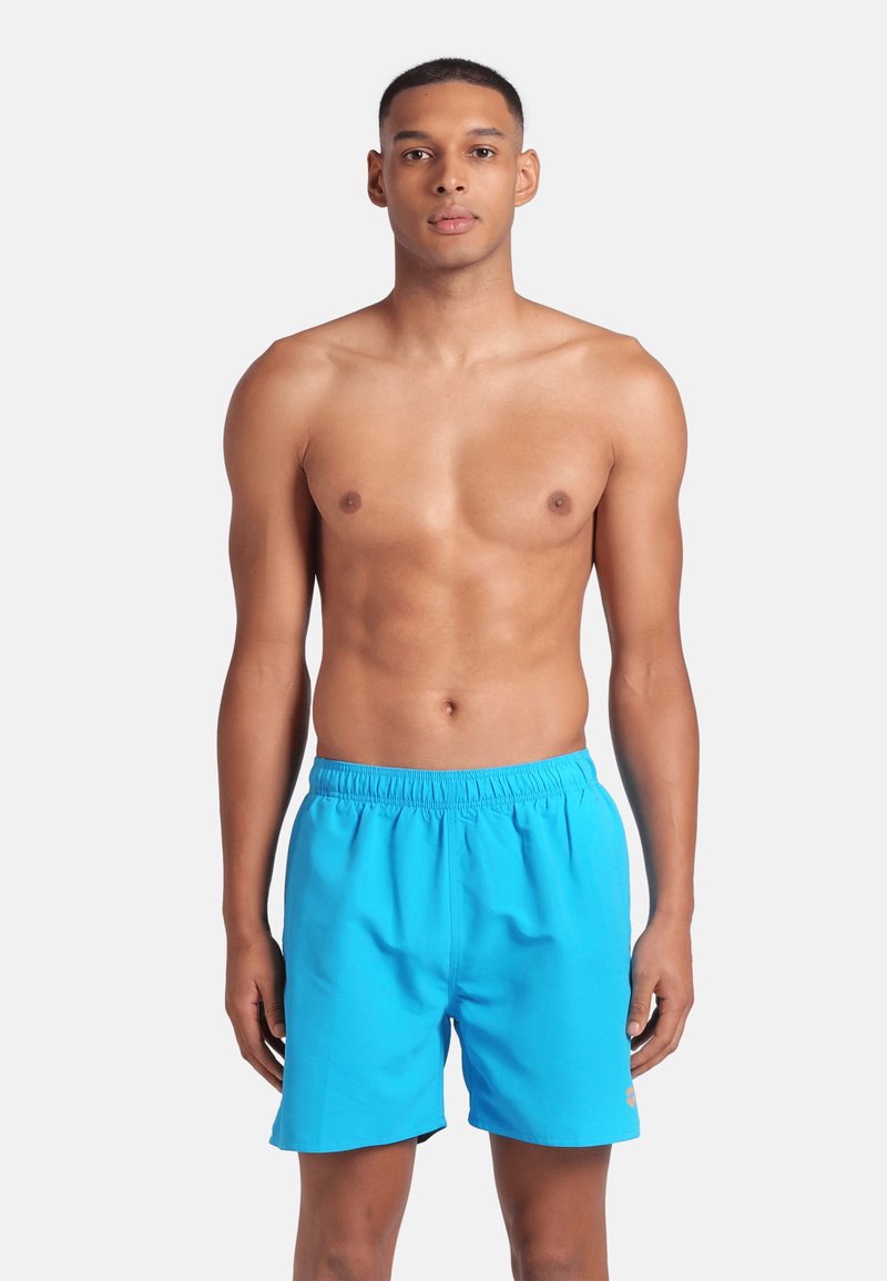 Modelo masculino de pie contra un fondo blanco, vistiendo bañadores azules brillantes con cintura elástica, mirando de frente.