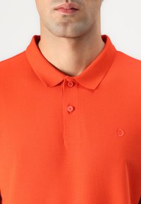 Camisa polo naranja con tejido texturizado, cuello y dos botones. Incluye un pequeño logo tonal en el lado izquierdo.