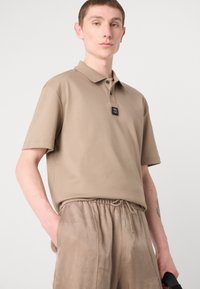 Korte mouw beige poloshirt met een kraag, zwart logo-label en twee knopen. Gekocht met lichtbruine, broek met koord.