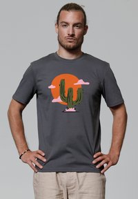 T-shirt en coton gris avec un graphisme de cactus vert portant une fleur rose, le tout devant un soleil orange et des nuages roses. Manches courtes et col rond.