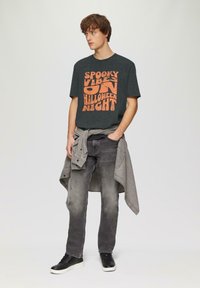 Zwarte t-shirt met oranje tekst die zegt "SPOOKY VIBES ON HALLOWEEN NIGHT," gecombineerd met grijze jeans en zwarte sneakers. Lichtgrijze shirt om de taille geknoopt.