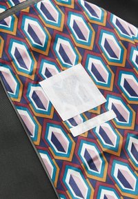 Mønstret stof med hexagonale design i lilla, blå, orange og creme, med en hvid lap og velcro-detaljer på en mørk baggrund.