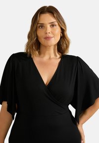Robe portefeuille noire avec des manches fluides, un décolleté en V et une taille cintrée. Le tissu est lisse et légèrement extensible, offrant du confort.
