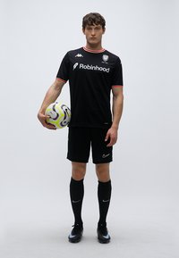 Maillot de football noir avec accents rouges, short noir assorti, chaussettes noires montantes jusqu'aux genoux et chaussures à crampons noires. Tenant un ballon de football multicolore.