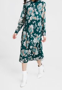 Sukienka z długim rękawem w ciemnozielonym materiale, zdobiona kwiecistym wzorem w odcieniach niebieskiego, kremowego i różowego, z zaznaczoną talią i swobodnym krojem.