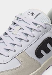 Λευκό και γκρι αθλητικό παπούτσι Etnies με λευκά κορδόνια και μαύρο λογότυπο στο πλάι, με έμφαση στις λεπτομέρειες του επάνω μέρους και της γλώσσας.