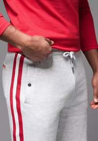 Pantalones de chándal grises con rayas rojas en los laterales, cinturilla elástica con cordón, bolsillos laterales y un detalle de botón negro. Parte superior de manga larga roja.