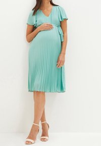 Mátově zelené plisované šaty s volutým výstřihem, rozšířenými krátkými rukávy a zavázaným pasem. Doplněno o světle páskové sandály.