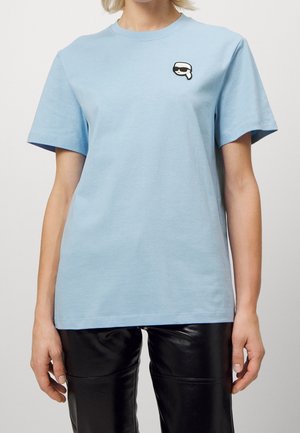 Personne portant un T-shirt bleu clair avec un petit patch représentant un visage avec des lunettes de soleil sur la poitrine gauche et un pantalon en cuir noir.