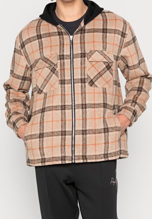 Kariertes Jackett in Beige, Orange und Schwarz. Mit einem Reißverschluss an der Vorderseite, zwei Brusttaschen und einer Kapuze aus schwarzem Stoff. Über einem schwarzen Hemd getragen.