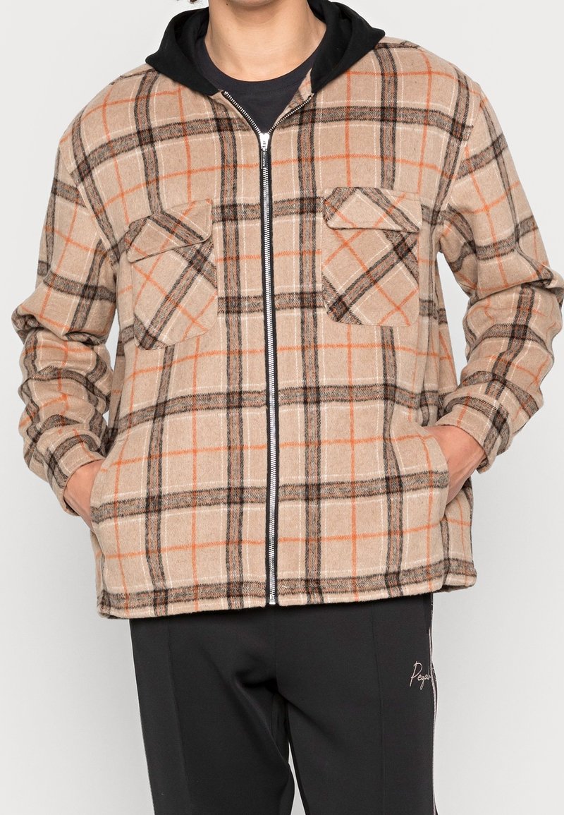 Rutig jacka i beige, orange och svart. Har dragkedja framtill, två bröstfickor och en huva i svart tyg. Bärs över en svart skjorta.