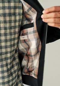 Sort jakkes indre har et lyst ternet for i beige, brunt og blåt. Ydersiden har et grønt og beige rutemønster.
