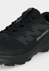 Μαύρο παπούτσι Salomon για τρέξιμο σε μονοπάτια με κορδόνια, υφασμάτινο επάνω μέρος με υφή και λογότυπα στο πλάι και στη γλώσσα.